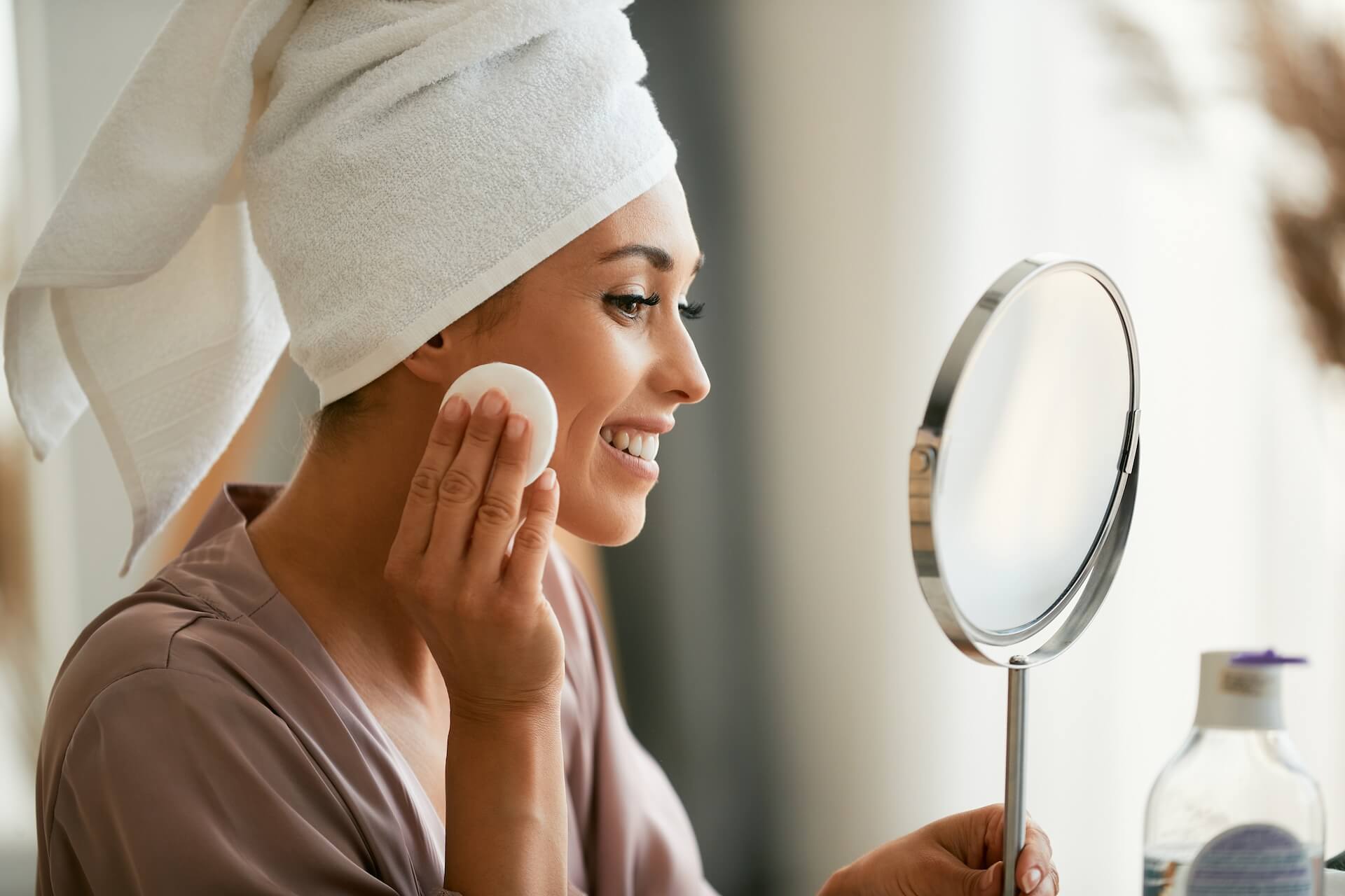 Young beautiful woman removing make-up of her face while looking herself in a mirror at home